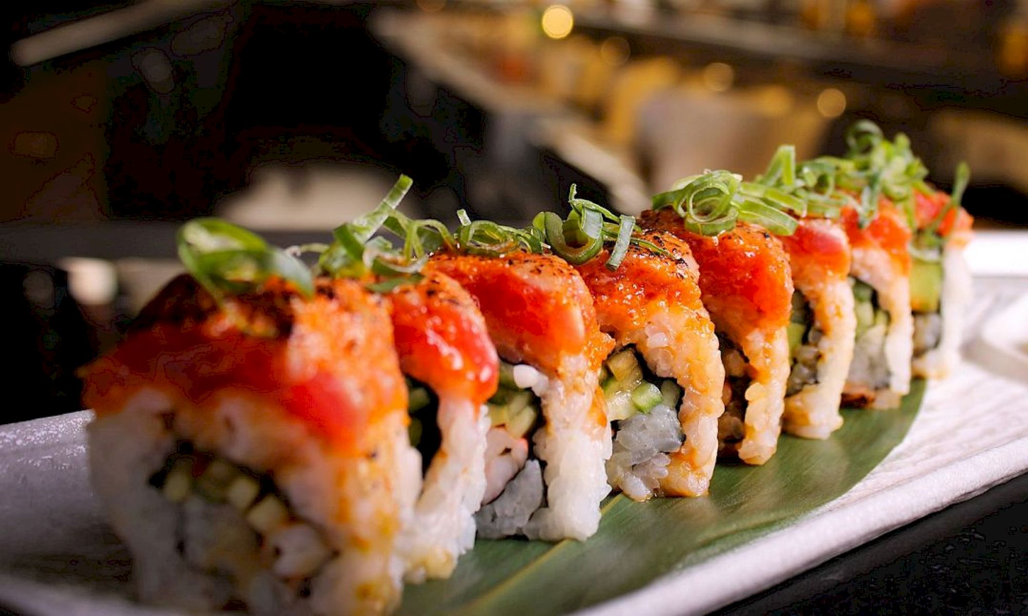 The image shows a sushi roll topped with a red sauce and green garnishes, neatly arranged on a leaf-lined plate in a restaurant setting.