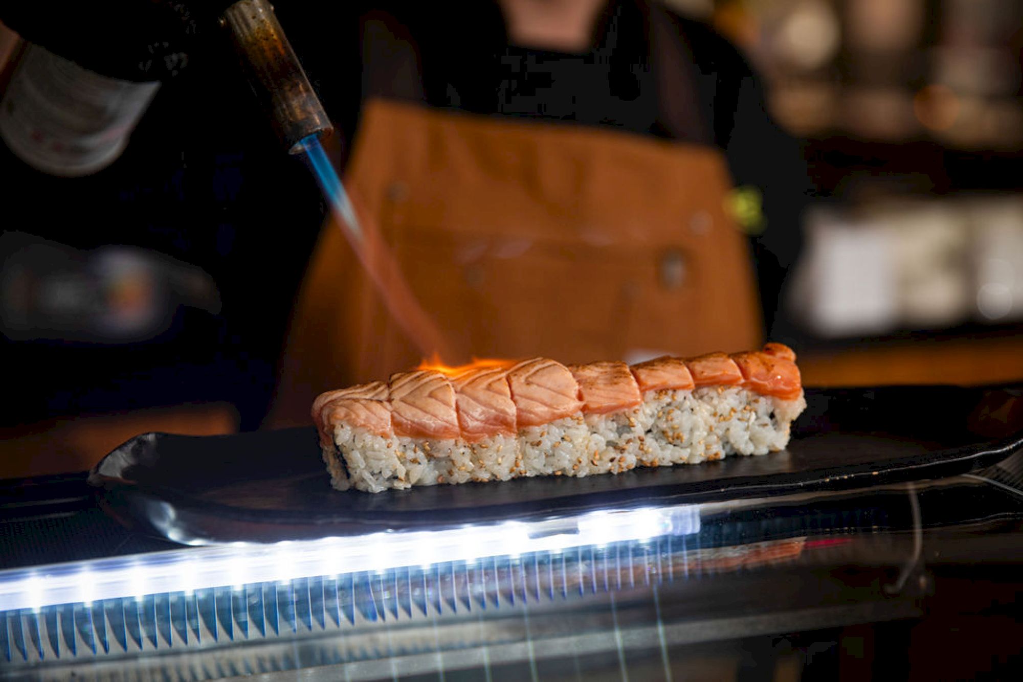A chef is using a blowtorch to sear a row of sushi, likely salmon, adding a finishing touch to the dish.