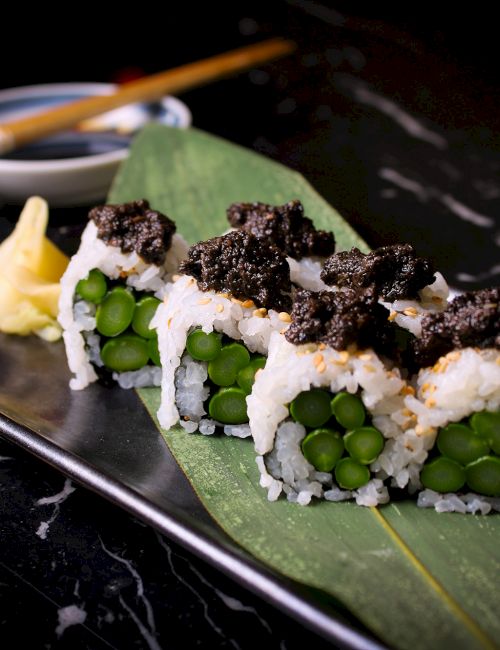 The image shows sushi rolls with asparagus and a dark topping, served on a leaf with ginger, wasabi, and soy sauce.