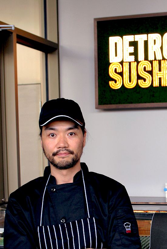 A person in chef attire stands in front of a sign that says "Detroit Sushi" inside a restaurant setting.