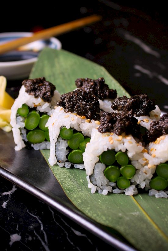 Sushi roll with asparagus, topped with black caviar, on a leaf. Served with wasabi, ginger, and soy sauce in the background.