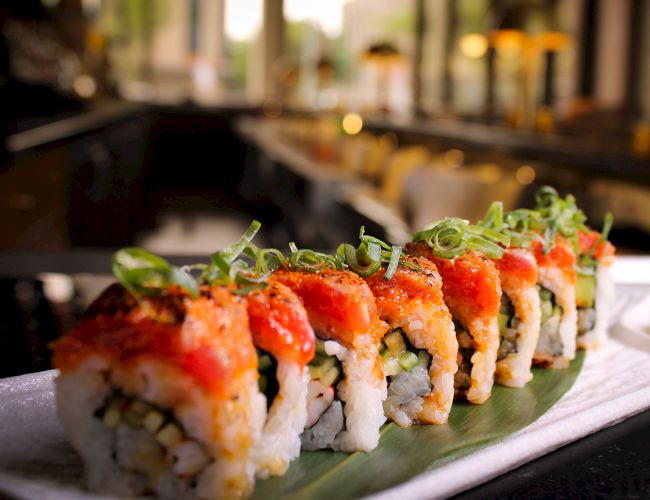 A plate of sushi rolls is garnished with green onions and features a vibrant red topping, served on a leaf with a blurred background setting.