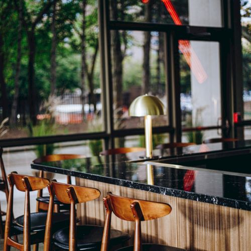 A modern bar setting with wooden chairs, a sleek black counter, a gold lamp, and large windows showing trees outside.