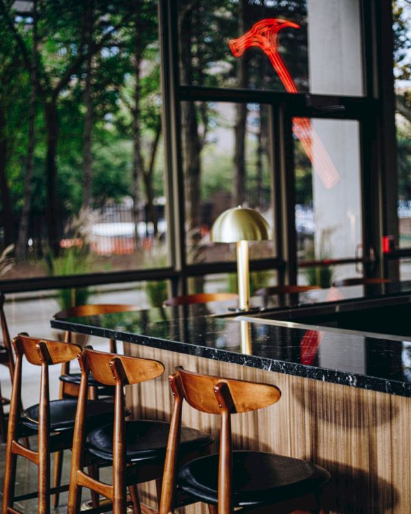 A modern bar setting with wooden chairs, a sleek black counter, a gold lamp, and large windows showing trees outside.