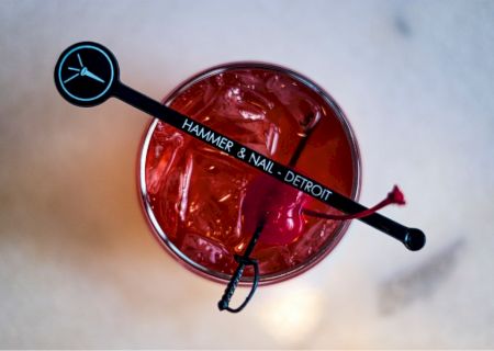 A red drink with ice sits in a glass, topped with a black stirrer labeled "Hammer & Nail - Detroit.".