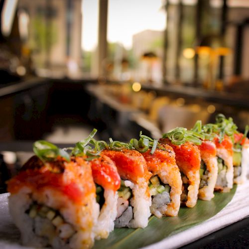 The image shows a sushi roll garnished with green onions, arranged on a leaf-covered plate in a restaurant setting.