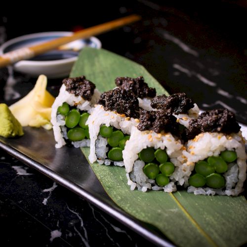 Vegetable sushi rolls with a green filling, topped with a dark garnish, served with wasabi, ginger, and soy sauce in a dish.
