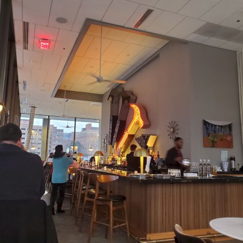 The image shows a modern bar with seating, decor, and patrons. Natural light comes through large windows, creating a cozy atmosphere.