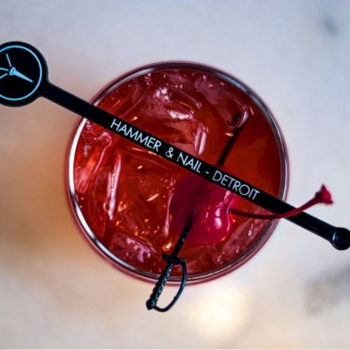 A red drink with ice and a stirrer labeled "HAMMER & NAIL - DETROIT" on a light surface.