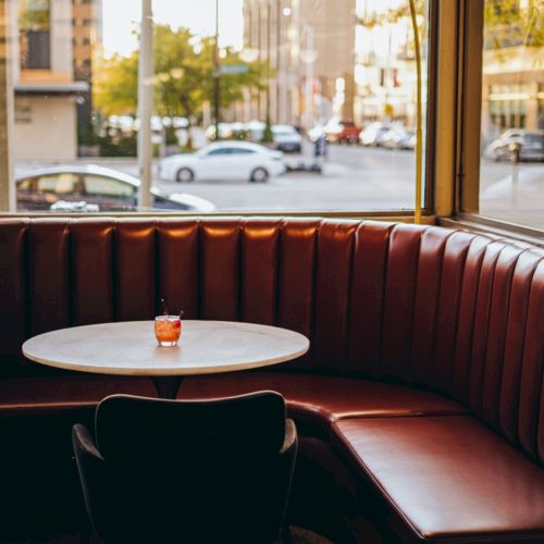 A cozy diner corner with a round table, single chair, and red booth, illuminated by a hanging lamp, with a city view outside the window.