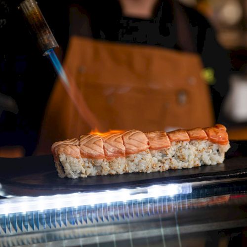 A chef is using a torch to sear the top of a sushi roll with slices of salmon, creating a grilled effect.
