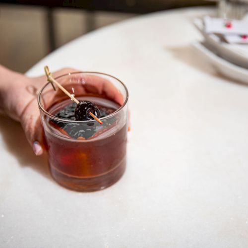 A hand holding a cocktail glass with a dark drink, garnished with a cherry on a toothpick, on a white table near plates and napkins.
