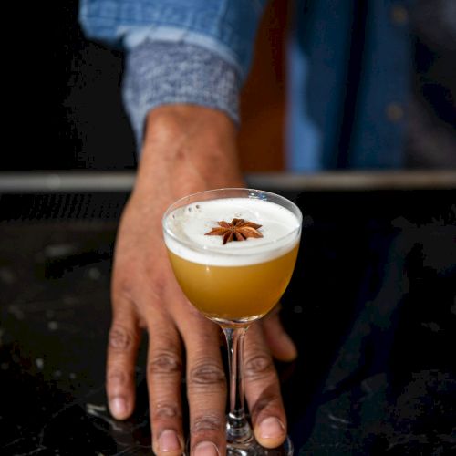 A person's hand is holding a cocktail glass with a frothy drink, garnished with a star anise on top.