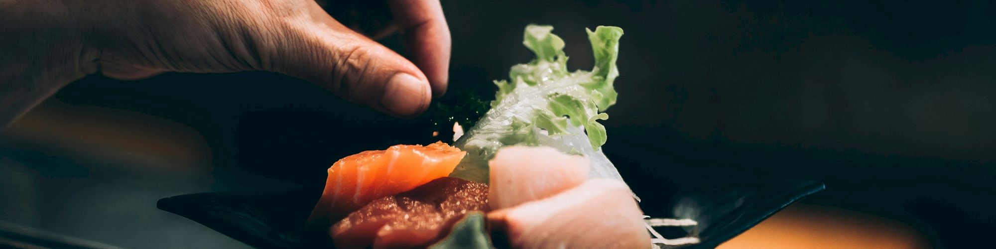A hand places sushi, including salmon and tuna slices, on a plate with lettuce garnish in an elegant setting.